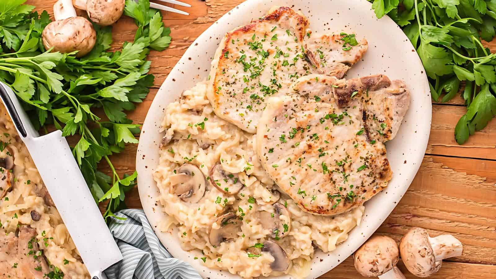 A plate with two grilled pork chops and creamy mushroom rice, garnished with chopped herbs. Fresh parsley and mushrooms are scattered around on a wooden table.