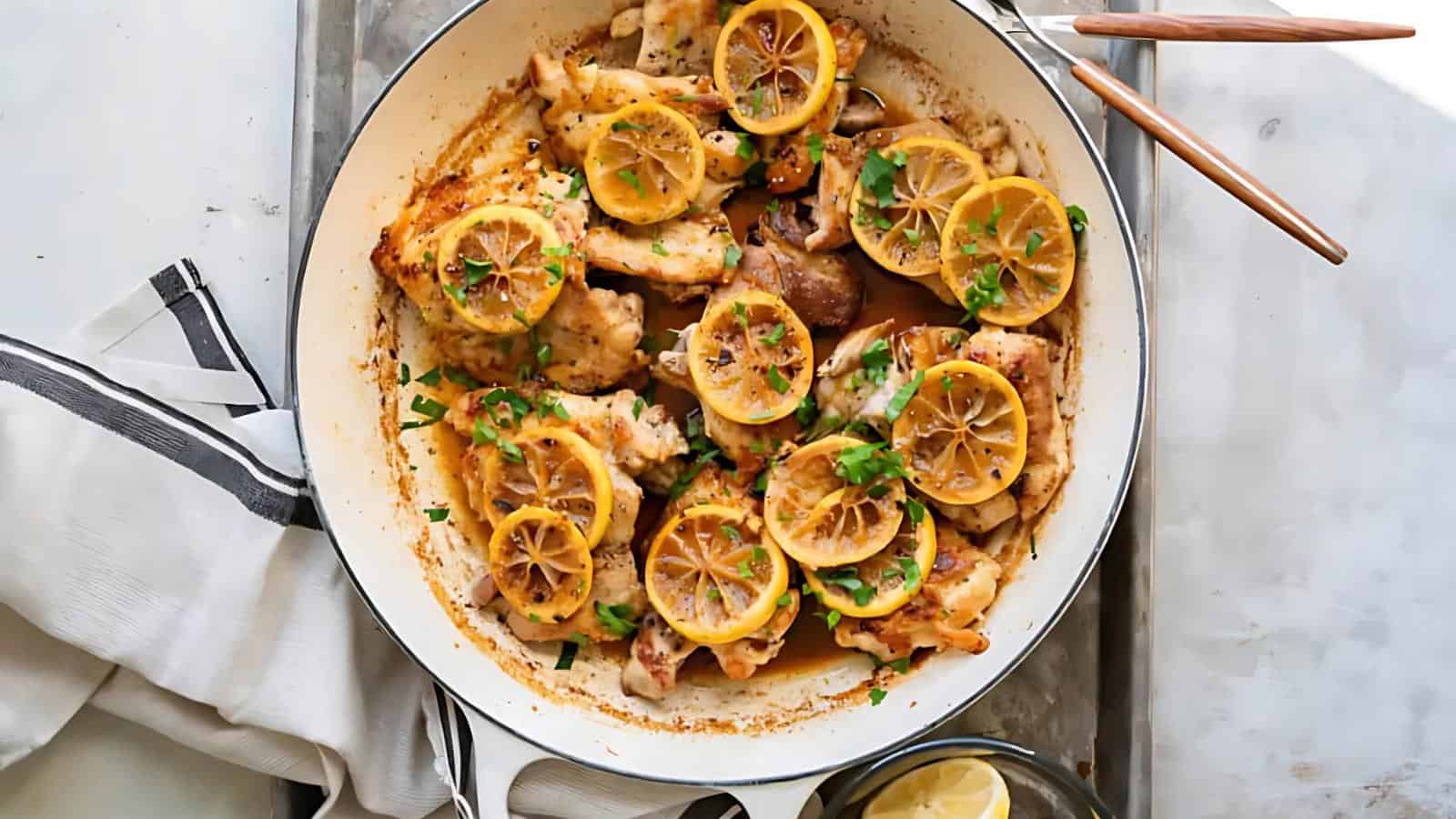 A skillet with cooked chicken pieces topped with lemon slices and chopped parsley, placed on a tray with a napkin and utensils nearby.