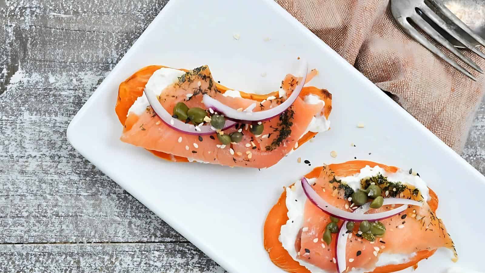 Two pieces of smoked salmon with cream cheese, capers, sesame seeds, and red onion slices on a white rectangular plate, next to a pink napkin and silver forks.