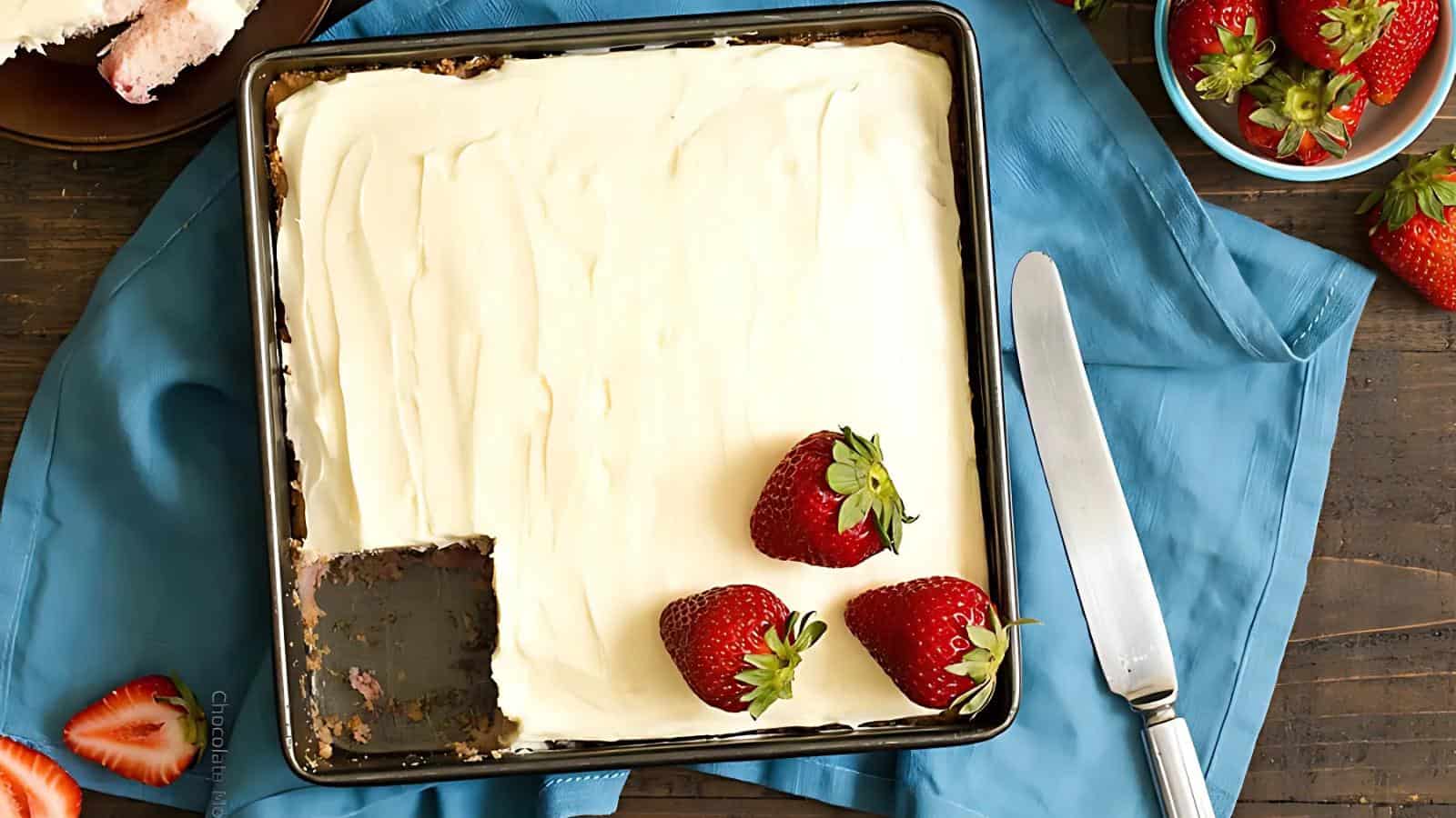 A square pan of frosted cake with one slice missing, topped with three whole strawberries, sits on a blue cloth next to a knife and a bowl of strawberries.