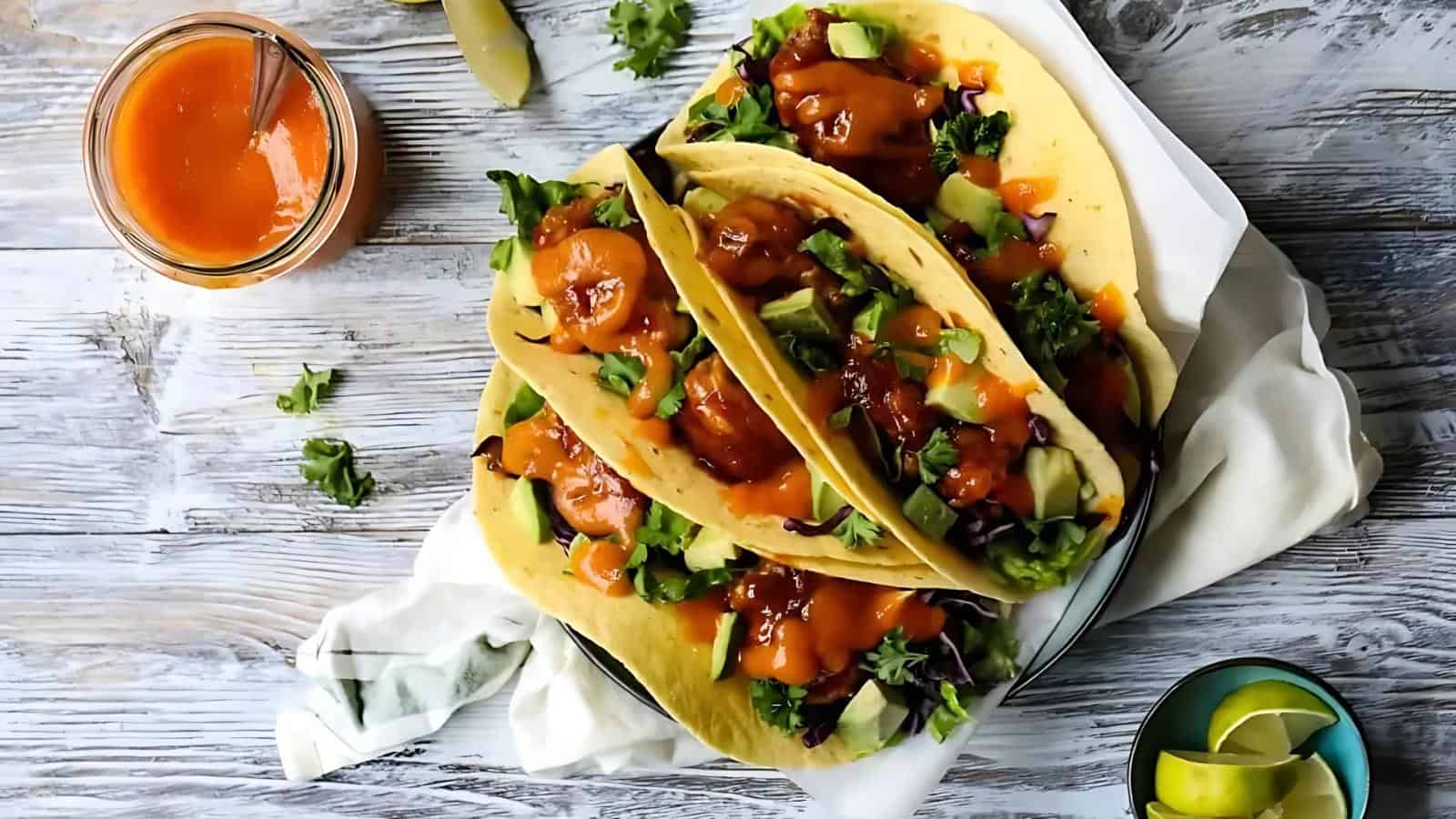 Three tacos filled with greens, avocado, and a red sauce on a plate. A jar of sauce and lime wedges are beside them on a wooden surface.
