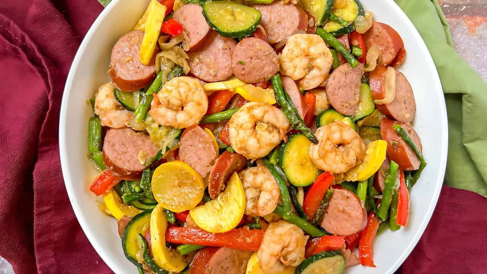 A white bowl filled with cooked shrimp, sausage slices, zucchini, bell peppers, and green beans.
