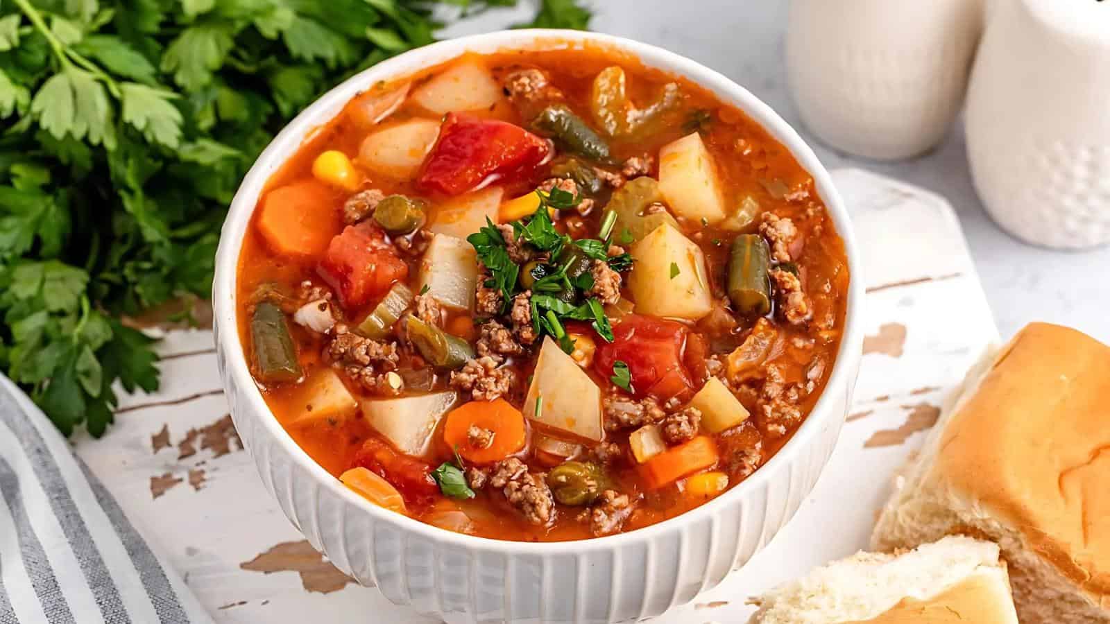 A bowl of vegetable beef soup with potatoes, carrots, tomatoes, green beans, corn, and ground beef. Garnished with parsley. Served with a dinner roll and parsley in the background.