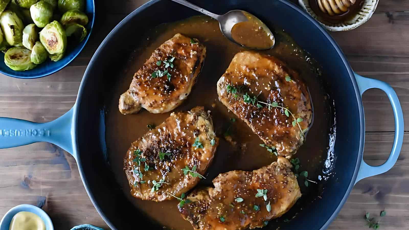 Four cooked pork chops with sauce and herbs in a blue skillet, with a serving spoon and a side of Brussels sprouts visible nearby.