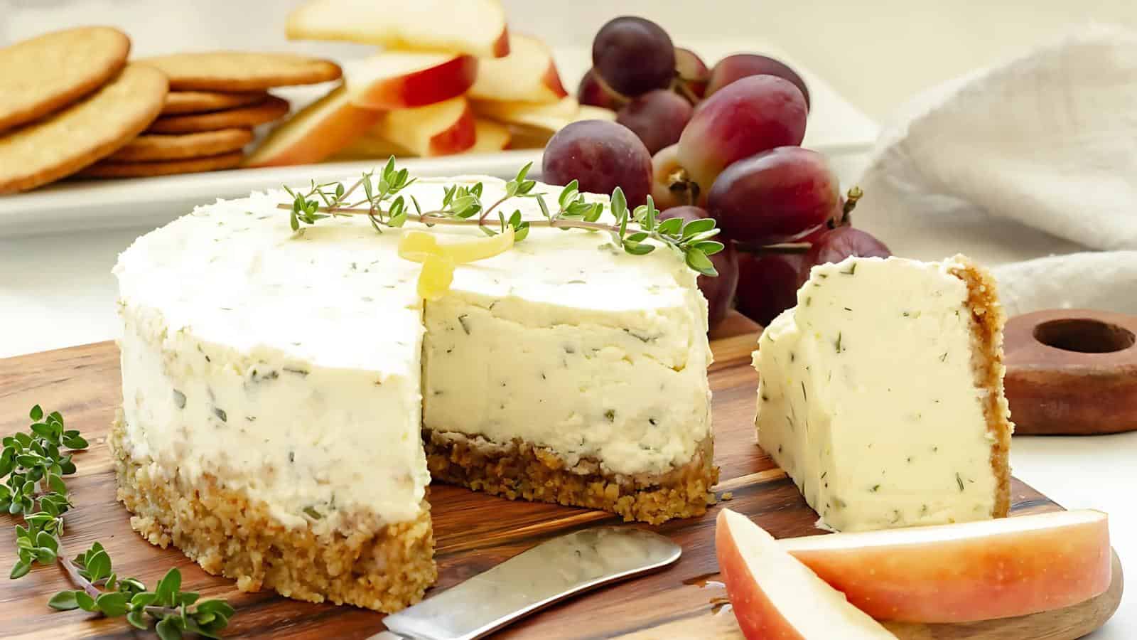 A round herb cheese wheel with a slice cut out, garnished with thyme, sits on a wooden board with apple slices and grapes in the background.