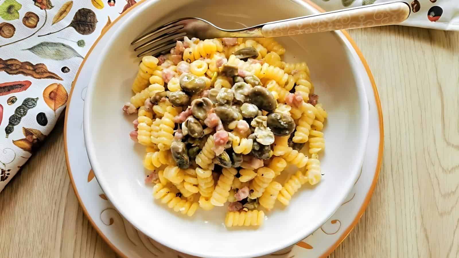 A plate of spiral pasta with a creamy sauce, diced ham, and green olives, served with a fork on a wooden table.
