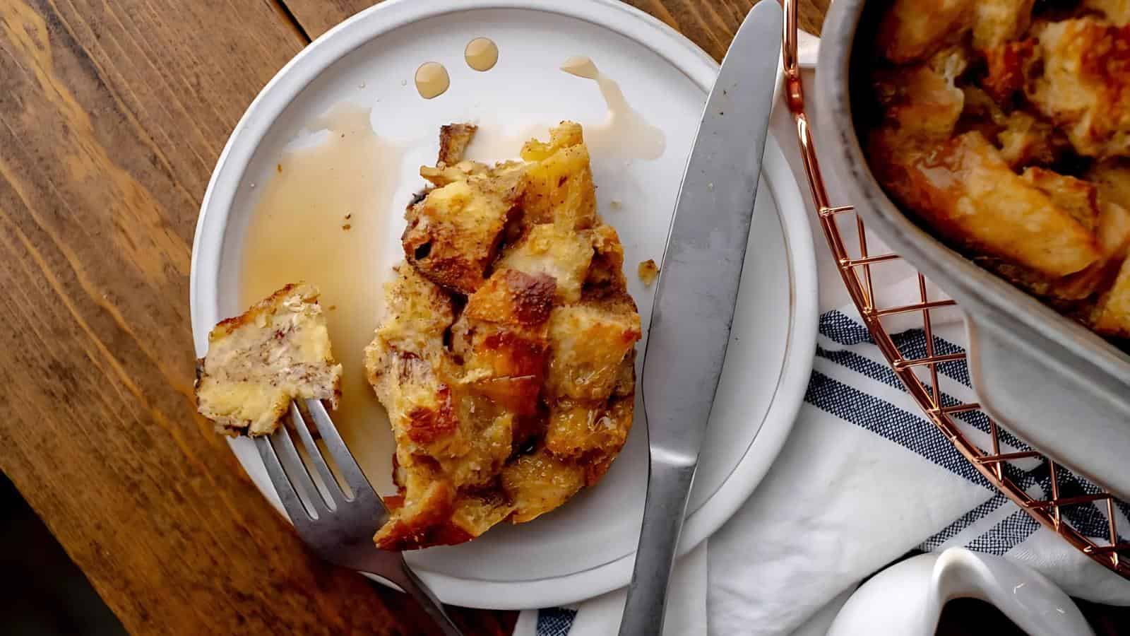 A slice of bread pudding on a white plate with a fork and knife, next to a pan of bread pudding. Syrup is drizzled over the dessert, and a striped cloth is nearby on a wooden table.