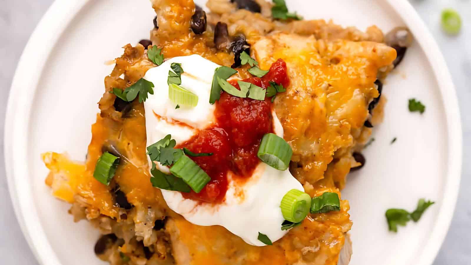 A serving of cheesy casserole topped with sour cream, salsa, chopped green onions, and cilantro on a white plate.