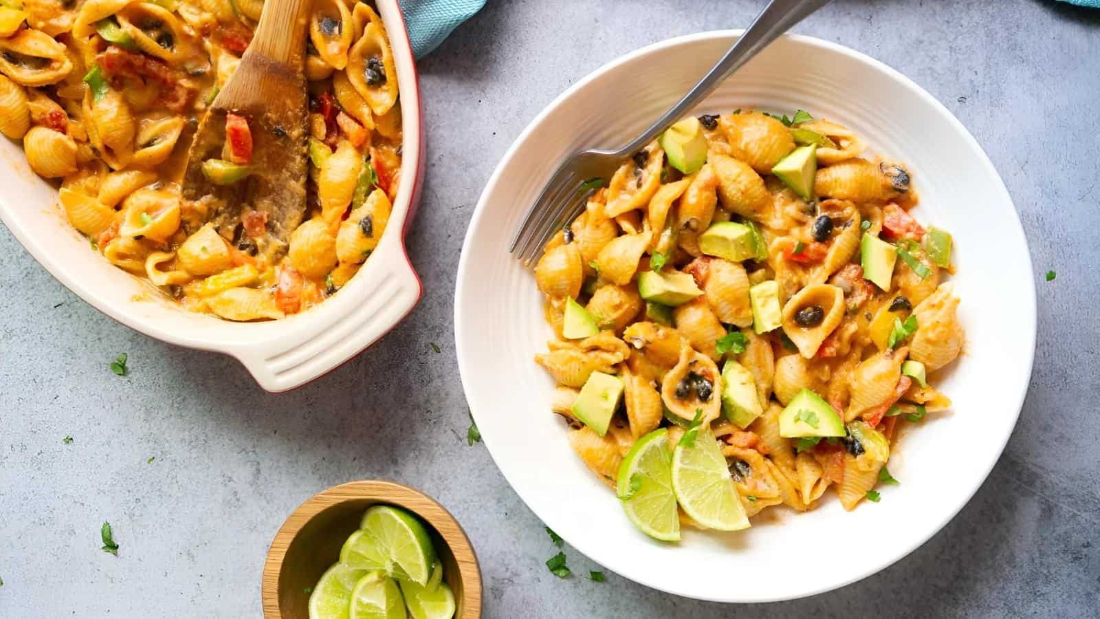 A plate of pasta shells with diced avocado and lime wedges. A serving dish with pasta and a wooden spoon is nearby, along with a small bowl of lime slices on a textured surface.
