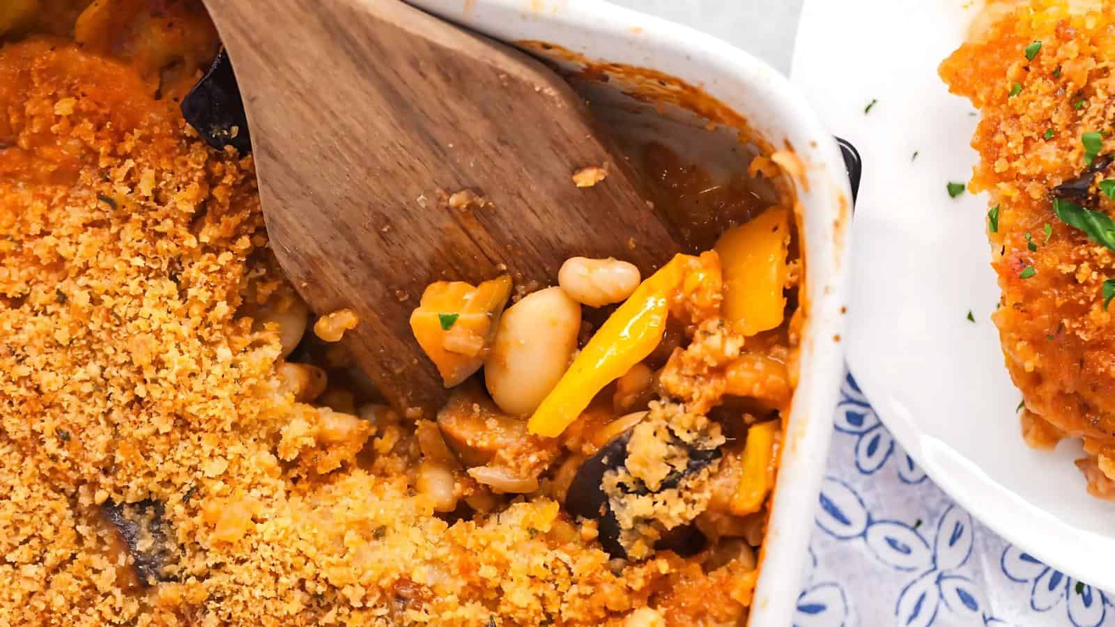 A close-up of a baked casserole topped with breadcrumbs, with white beans, yellow bell peppers, and a wooden spatula inside the dish.