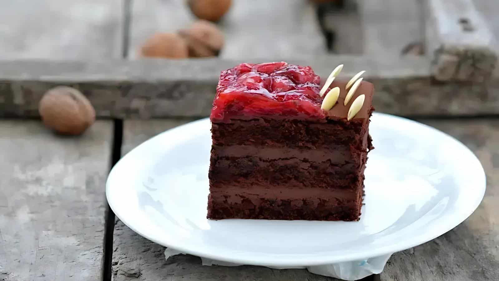 A slice of layered chocolate cake topped with a cherry glaze and almond slivers, served on a white plate on a rustic wooden surface.