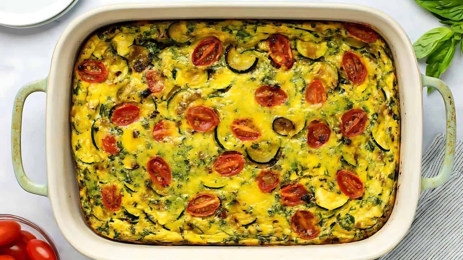 A baked vegetable frittata in a rectangular dish, topped with sliced cherry tomatoes, zucchini, and herbs.