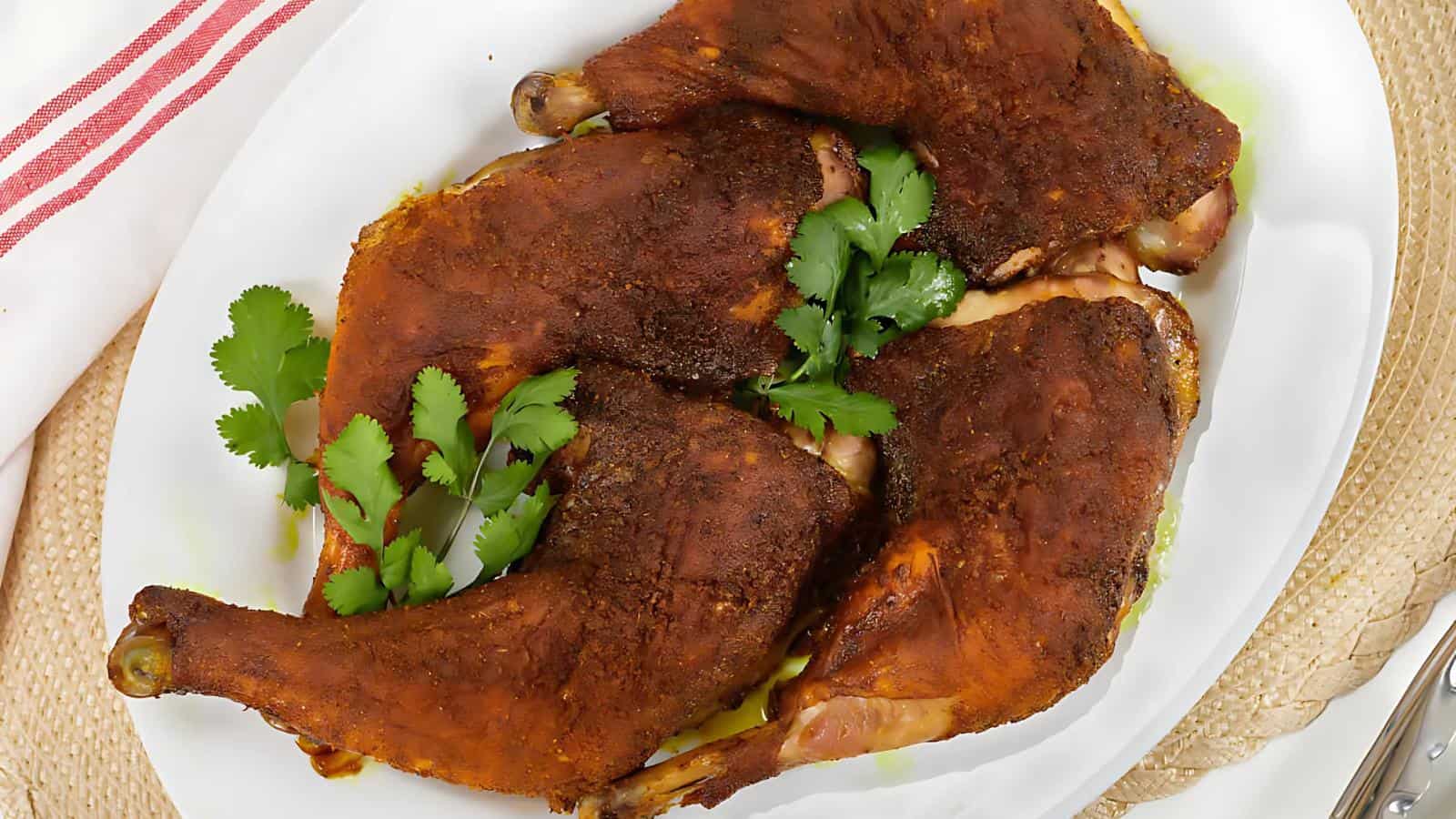 Four seasoned and roasted chicken leg quarters are arranged on a white plate, garnished with fresh cilantro.