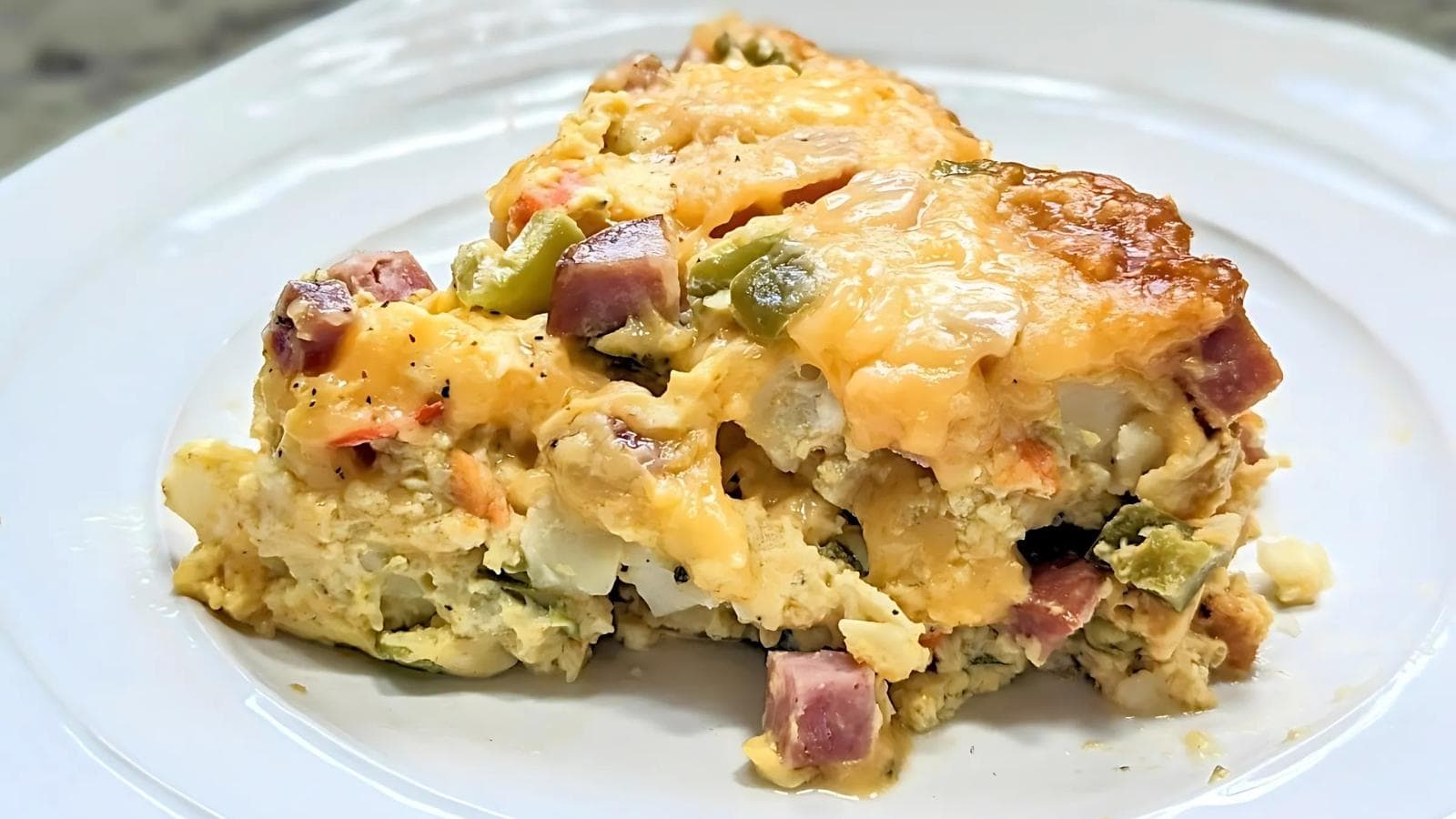 A piece of casserole with ham, cheese, and vegetables on a white plate.