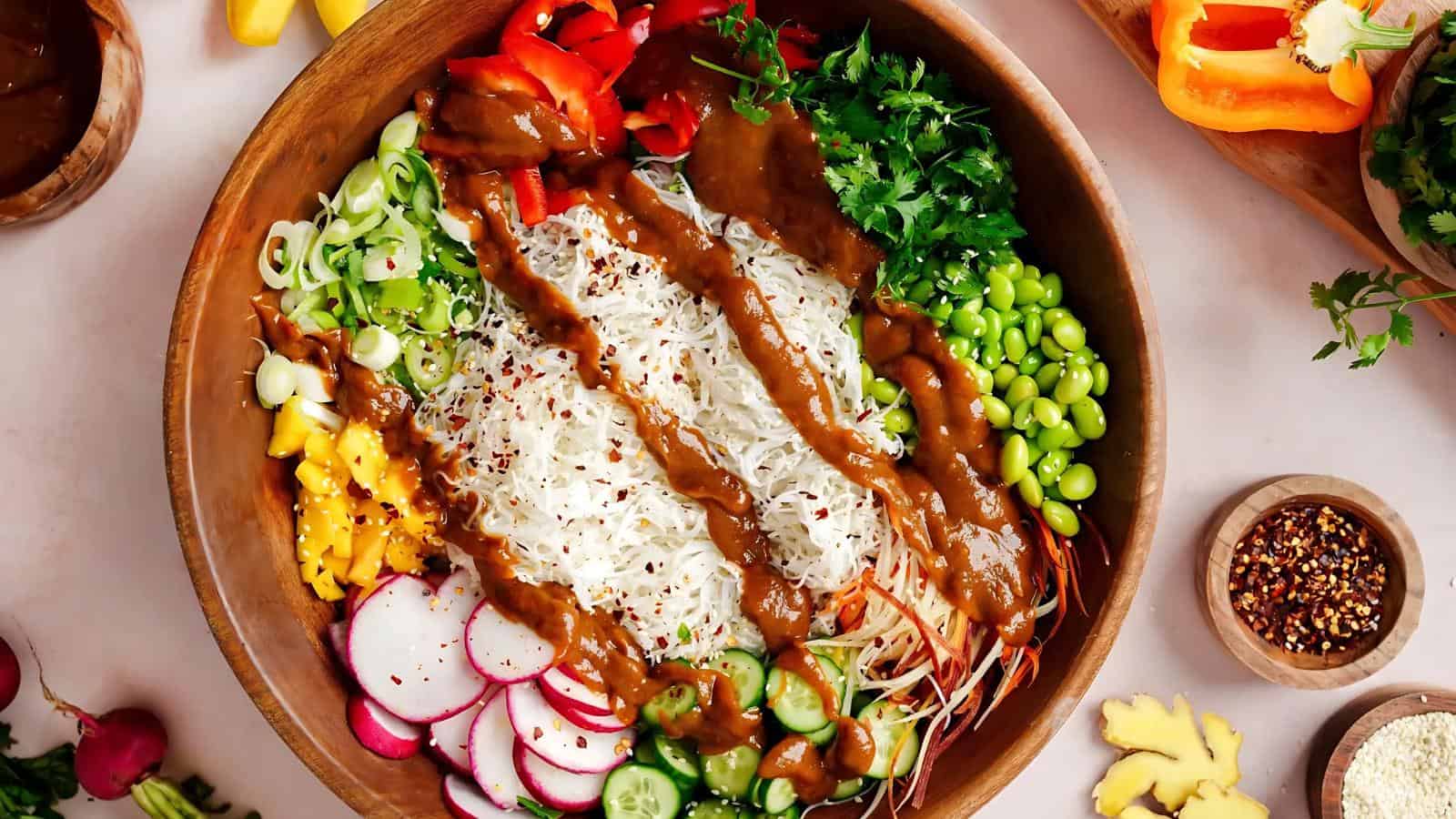 A wooden bowl filled with rice noodles, sliced vegetables, edamame, fresh herbs, and drizzled with brown sauce.