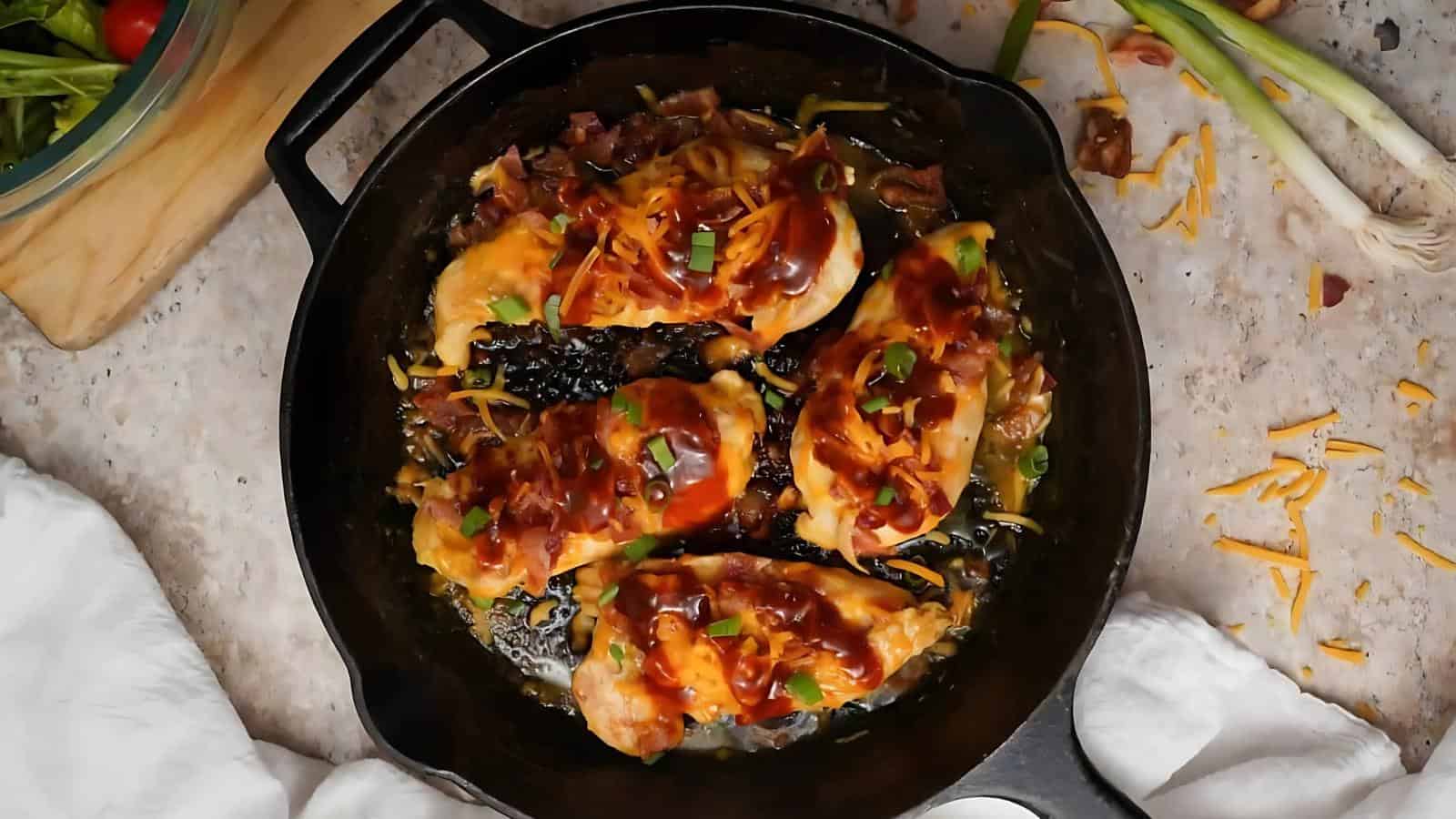 Four cooked chicken breasts topped with sauce, shredded cheese, and chopped green onions in a black cast iron skillet on a countertop.