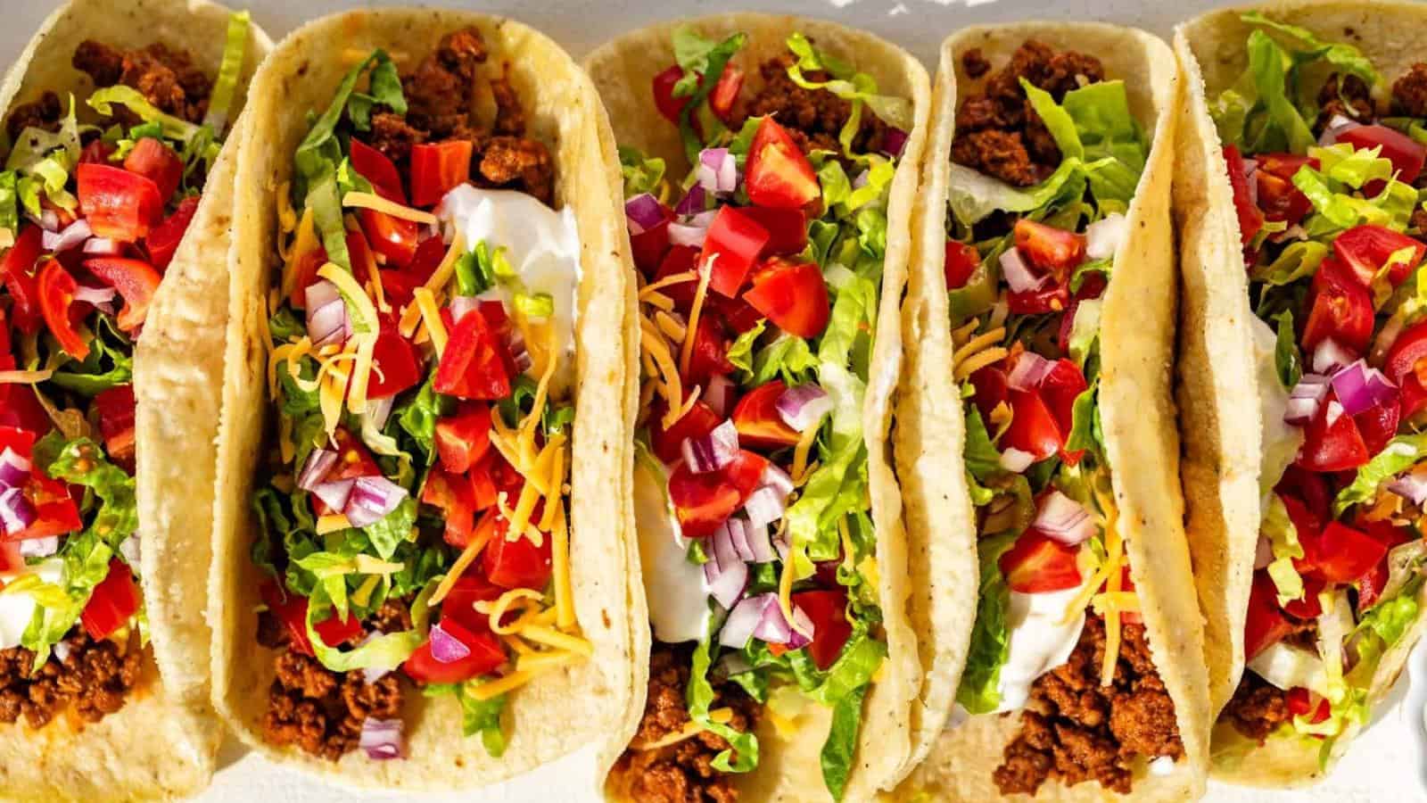 Five tacos filled with ground meat, shredded lettuce, diced tomatoes, chopped red onions, grated cheese, and sour cream, arranged side by side on a white surface.