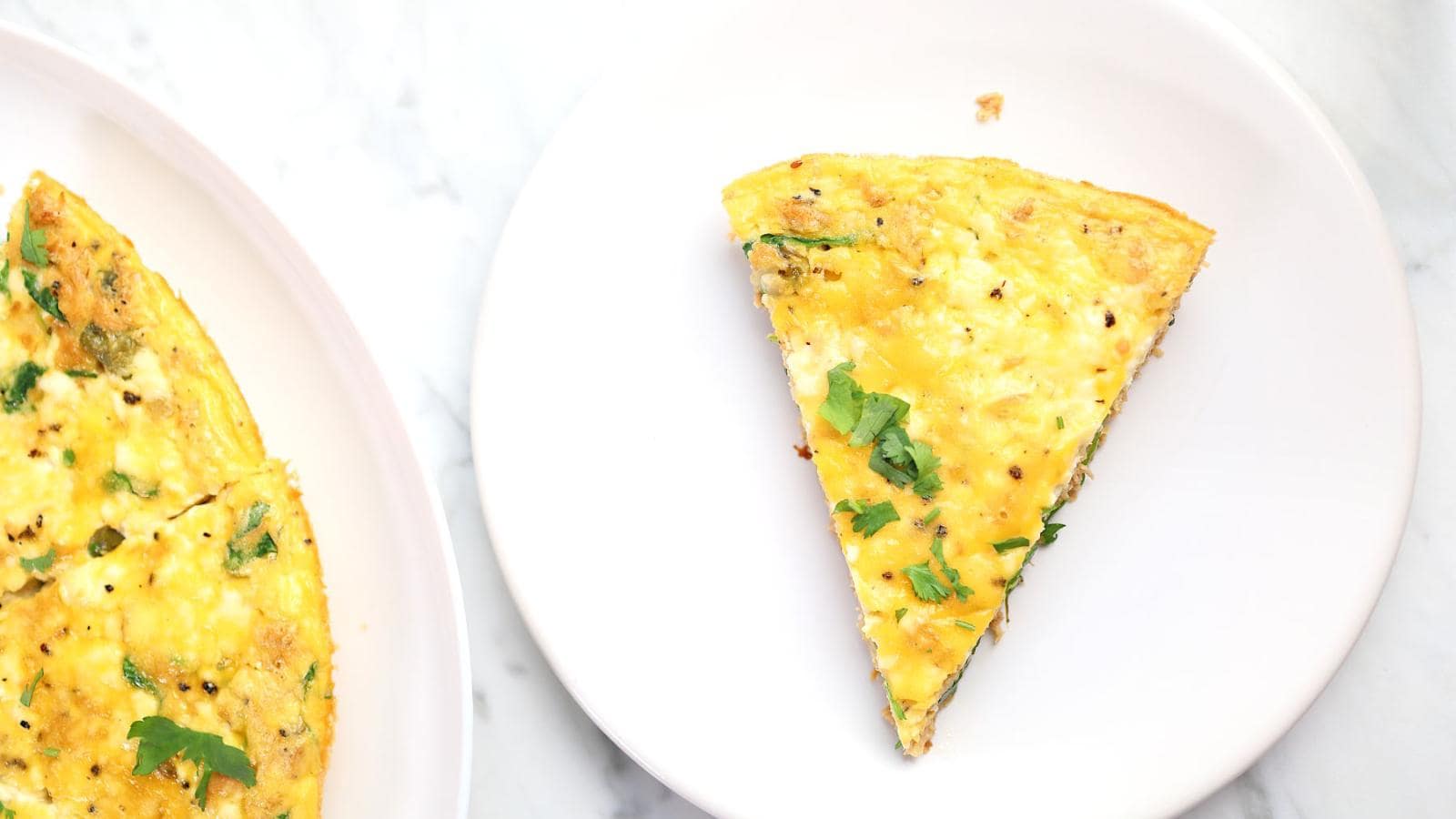A slice of vegetable frittata is served on a white plate next to the remaining frittata, both placed on a white surface.