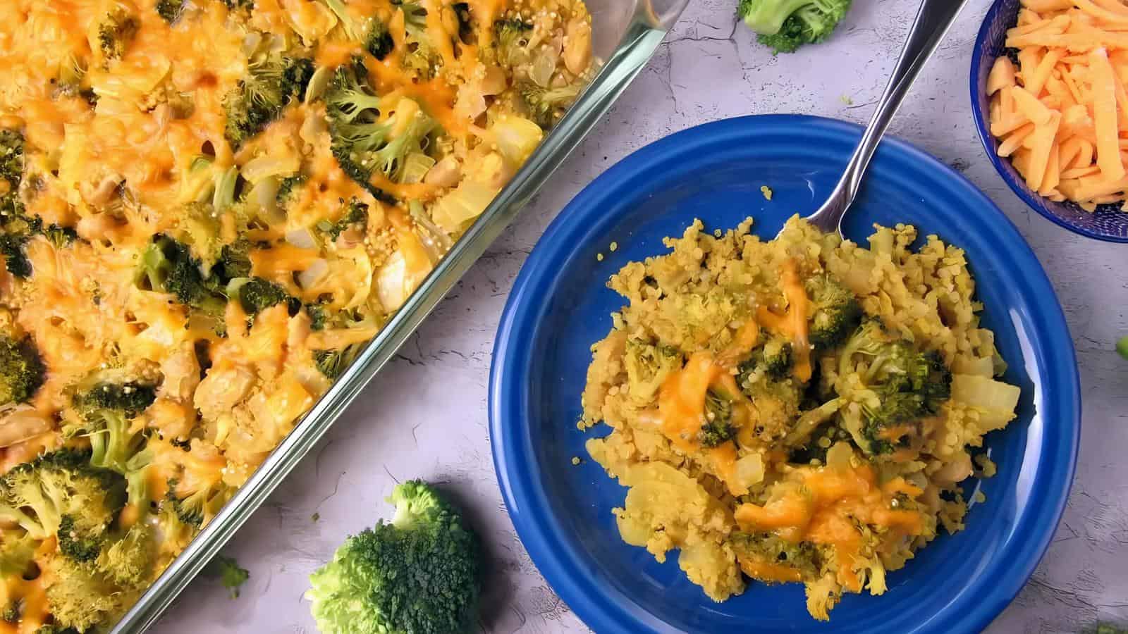 A serving of broccoli and cheese casserole on a blue plate, next to a glass baking dish with more casserole and a bowl of shredded cheese.