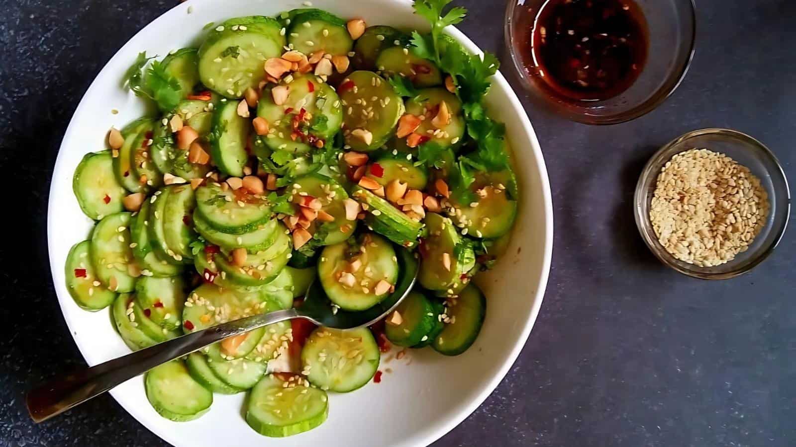A bowl of sliced cucumber salad topped with chopped peanuts and herbs, with a spoon, alongside small bowls of sauce and sesame seeds on a dark surface.