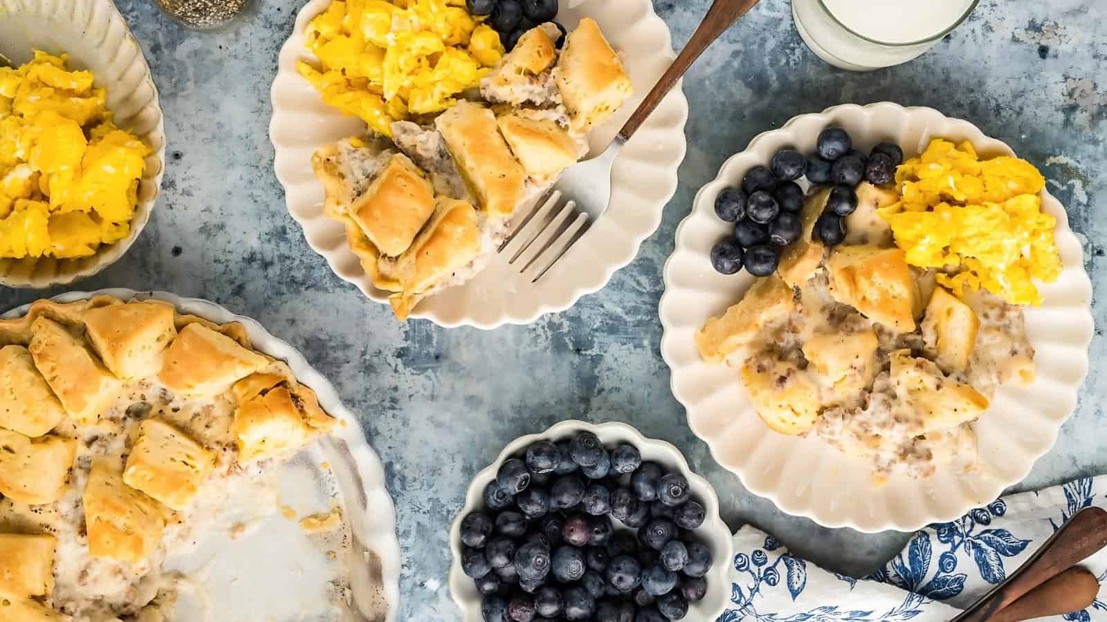 A table setting with plates of scrambled eggs, biscuits with gravy, and a bowl of blueberries. There's also a glass of milk, salt and pepper shakers, and a blue patterned napkin.