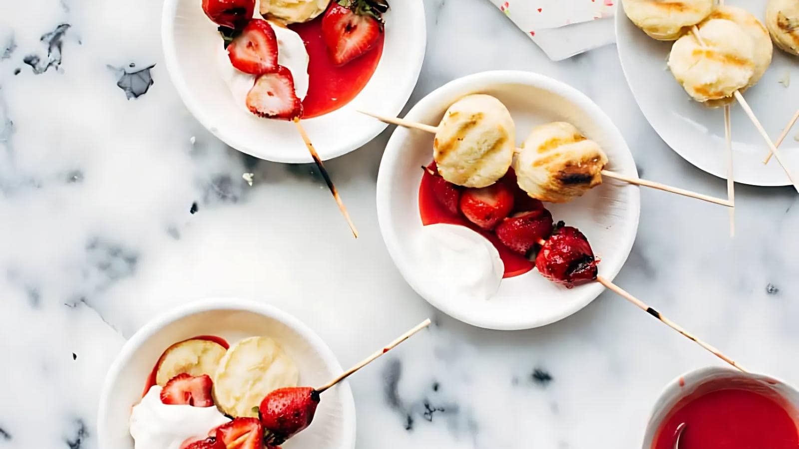 Plates with skewered grilled biscuits and strawberries, served with whipped cream and strawberry sauce on a marble surface.