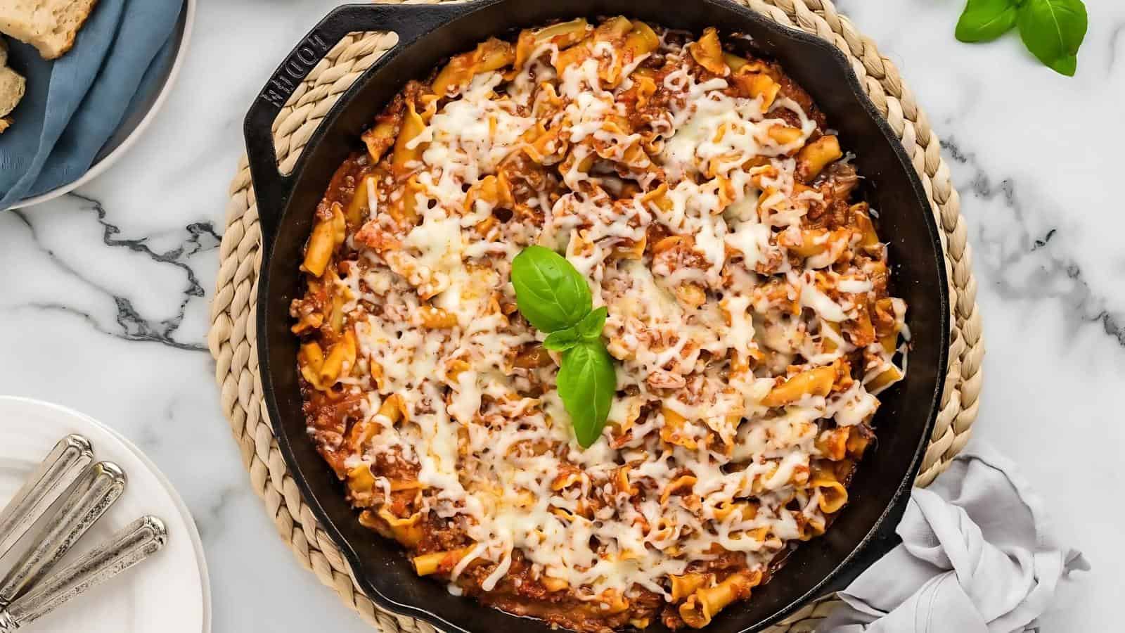 A skillet filled with baked pasta in tomato sauce, topped with melted cheese and garnished with a sprig of basil, sits on a woven trivet on a marble surface.