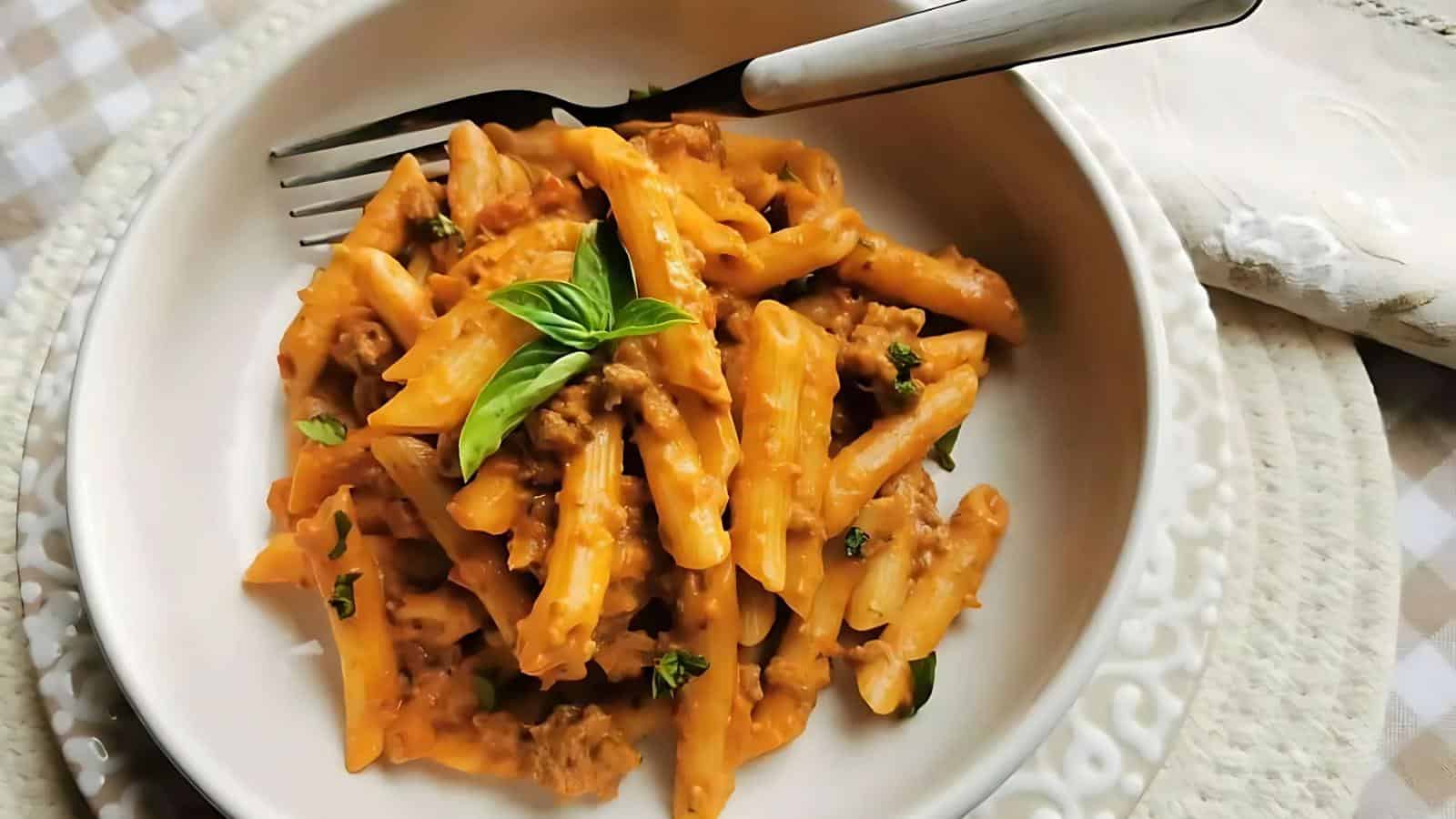 A bowl of penne pasta with creamy tomato sauce, ground meat, and fresh basil, served with a fork on a white patterned plate.