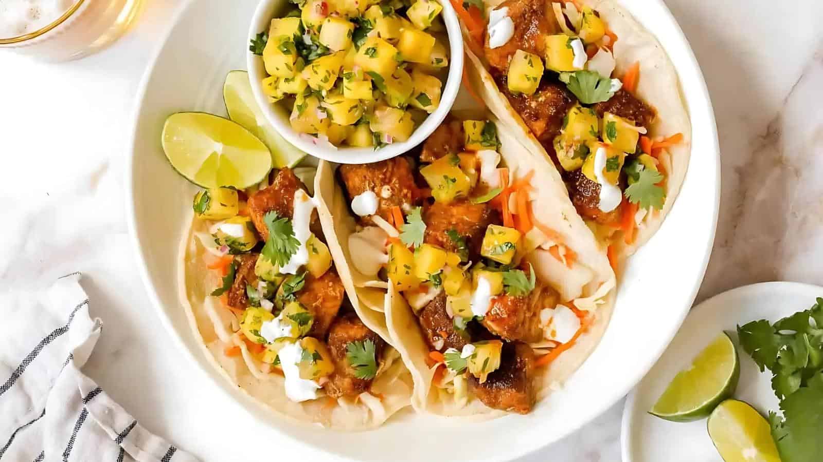 A plate with three tacos filled with meat, topped with pineapple salsa and cilantro, served with lime wedges and a small bowl of extra salsa.