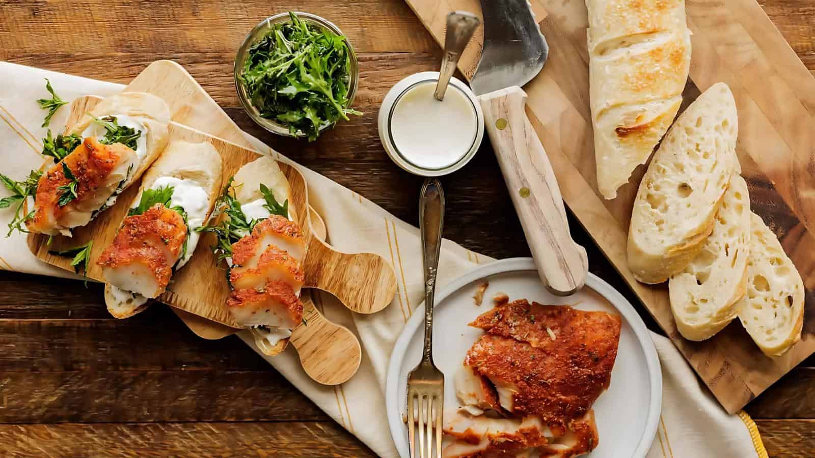 Sliced bread, pieces of seasoned fish, greens, and a small bowl of sauce are arranged on wooden boards and a plate atop a rustic wooden table.