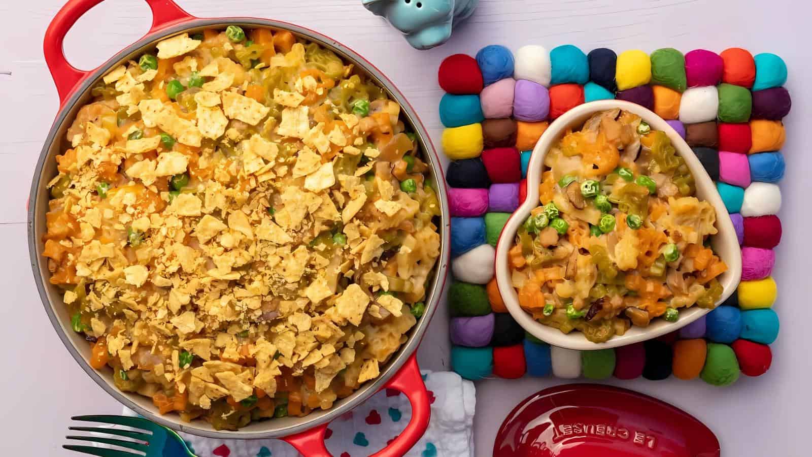 A casserole topped with crushed chips and a serving of the dish in a heart-shaped bowl, both on a white table with colorful accessories.