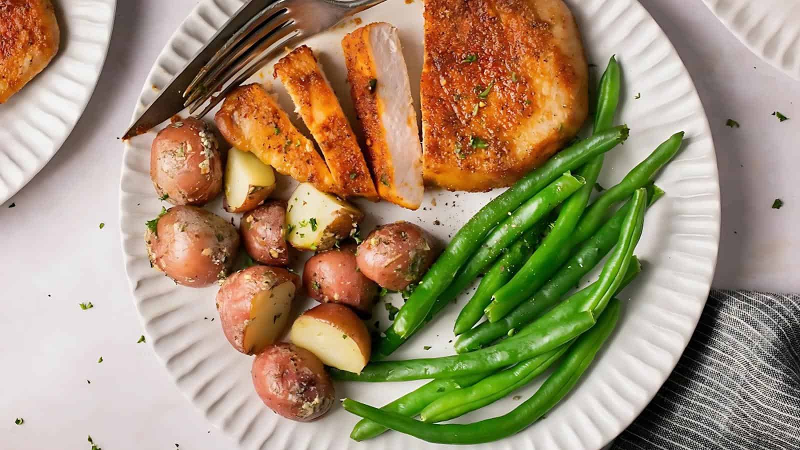 A plate with sliced seasoned pork chop, green beans, and halved red potatoes, with a fork and knife on the edge of the plate.