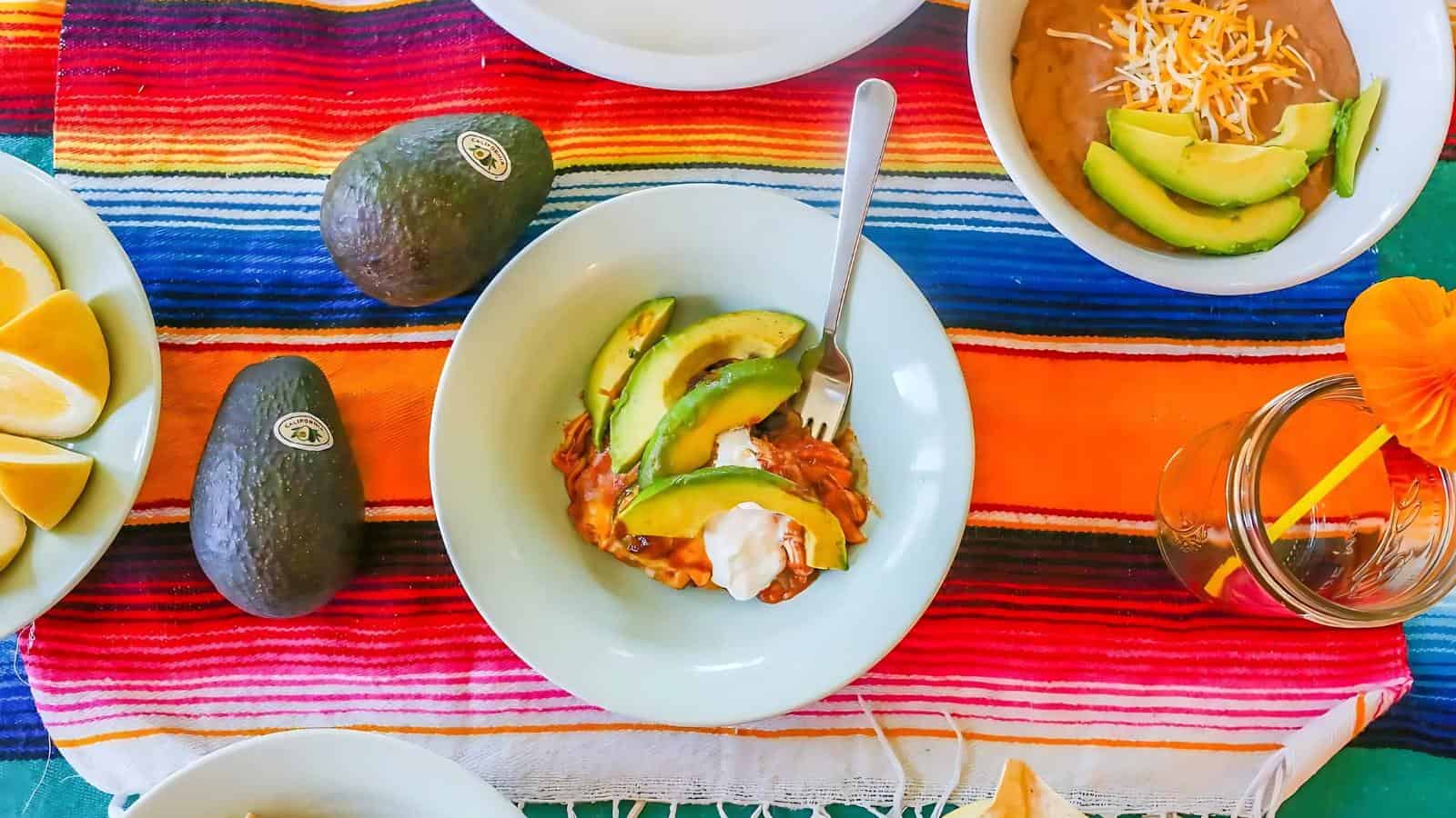 A table set with avocados, a bowl of beans topped with avocado and cheese, lemon wedges, and a plate of food with avocado and sour cream on a colorful striped tablecloth.