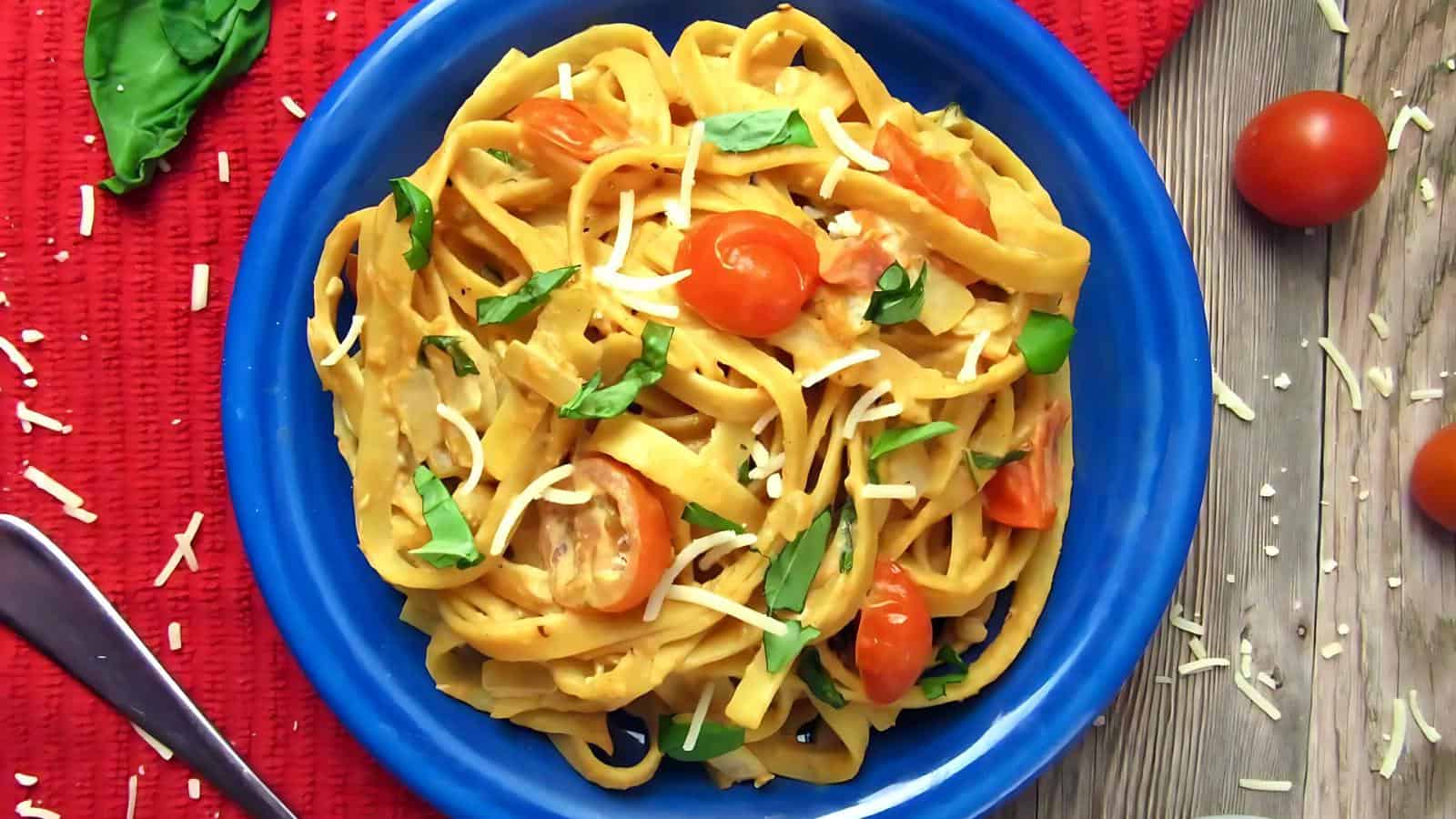 A blue plate filled with fettuccine pasta, cherry tomatoes, shredded cheese, and fresh basil, placed on a red textured cloth.