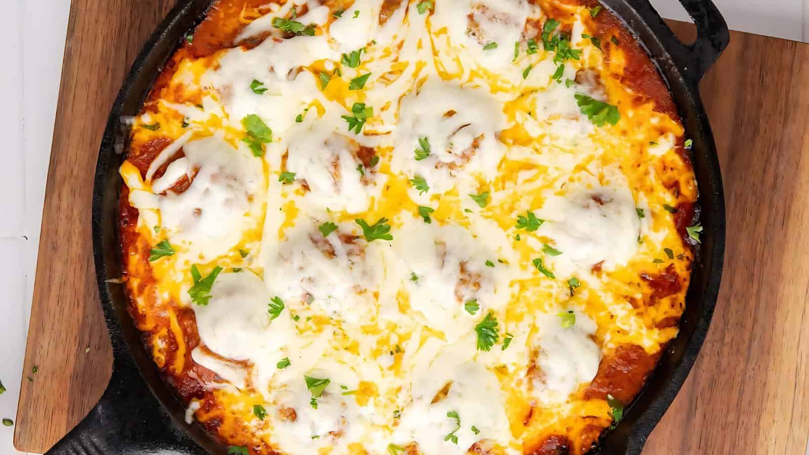 A skillet filled with baked meatballs topped with melted cheese and garnished with chopped parsley, placed on a wooden board.