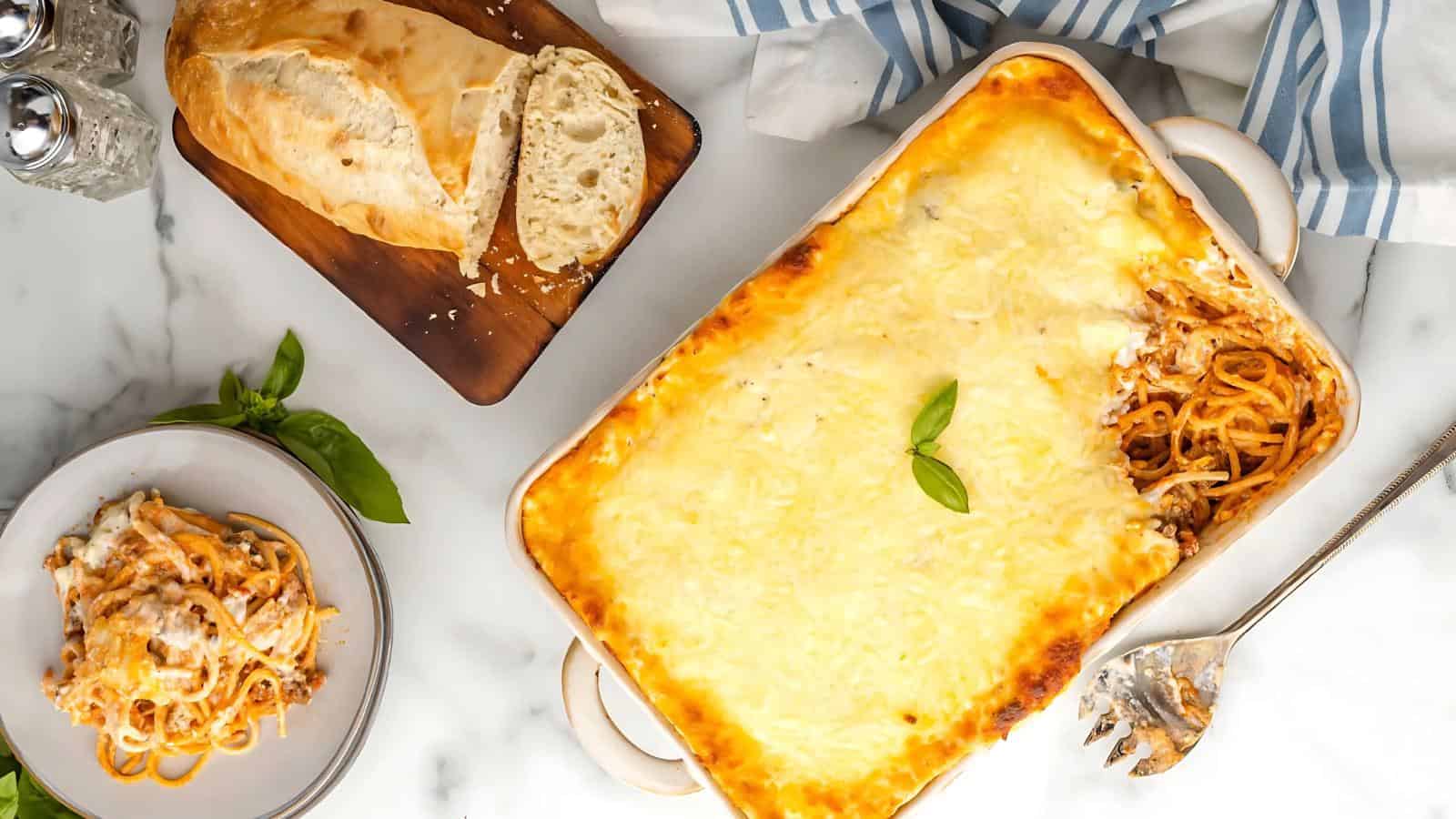 A baked pasta dish with a slice missing, topped with cheese and garnished with basil. Nearby are a cutting board with sliced bread and a salt and pepper set on a marble surface.