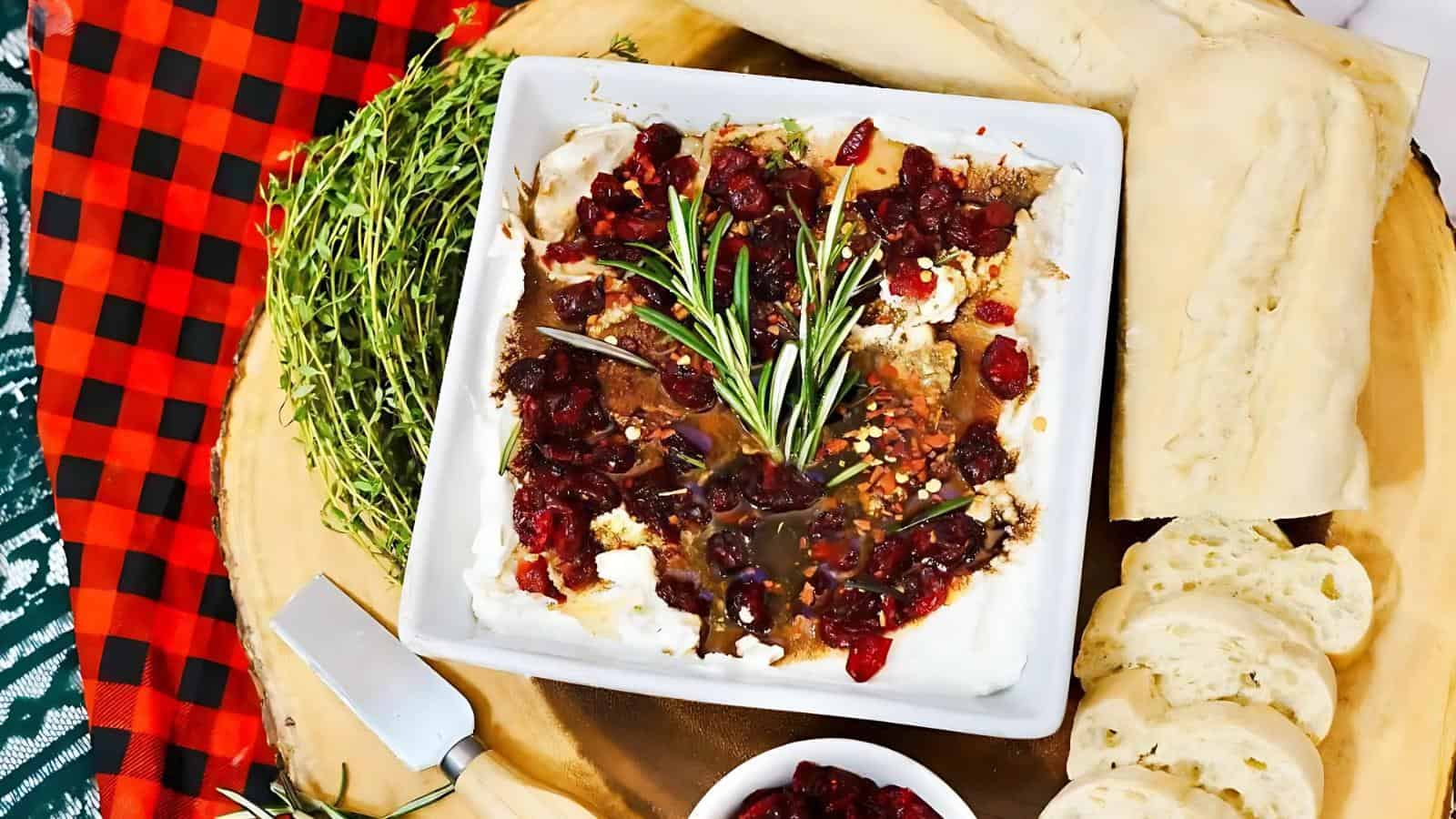Square dish of whipped cheese topped with cranberries and herbs, served on a wooden board with fresh bread, a cheese knife, and a red and black checkered cloth.
