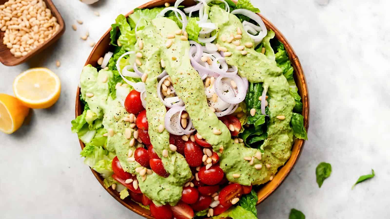 A salad with cherry tomatoes, lettuce, sliced onions, pine nuts, and green dressing in a wooden bowl, with lemon halves and a bowl of pine nuts nearby.