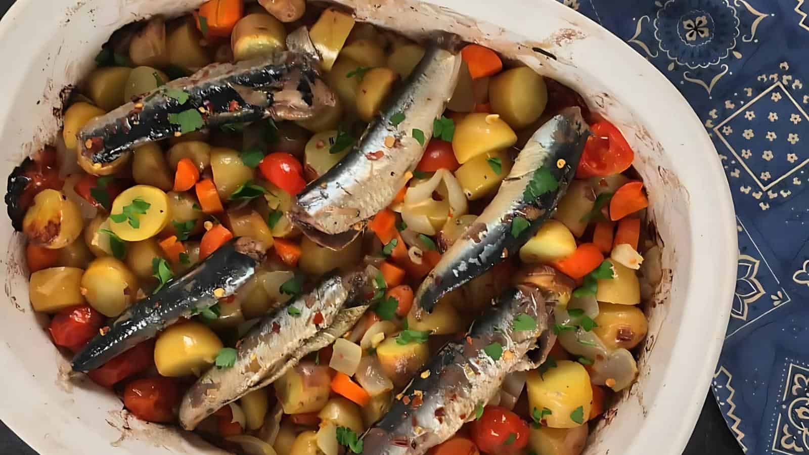 A baked dish with whole sardines, halved potatoes, cherry tomatoes, carrots, onions, and fresh parsley in a white casserole dish.
