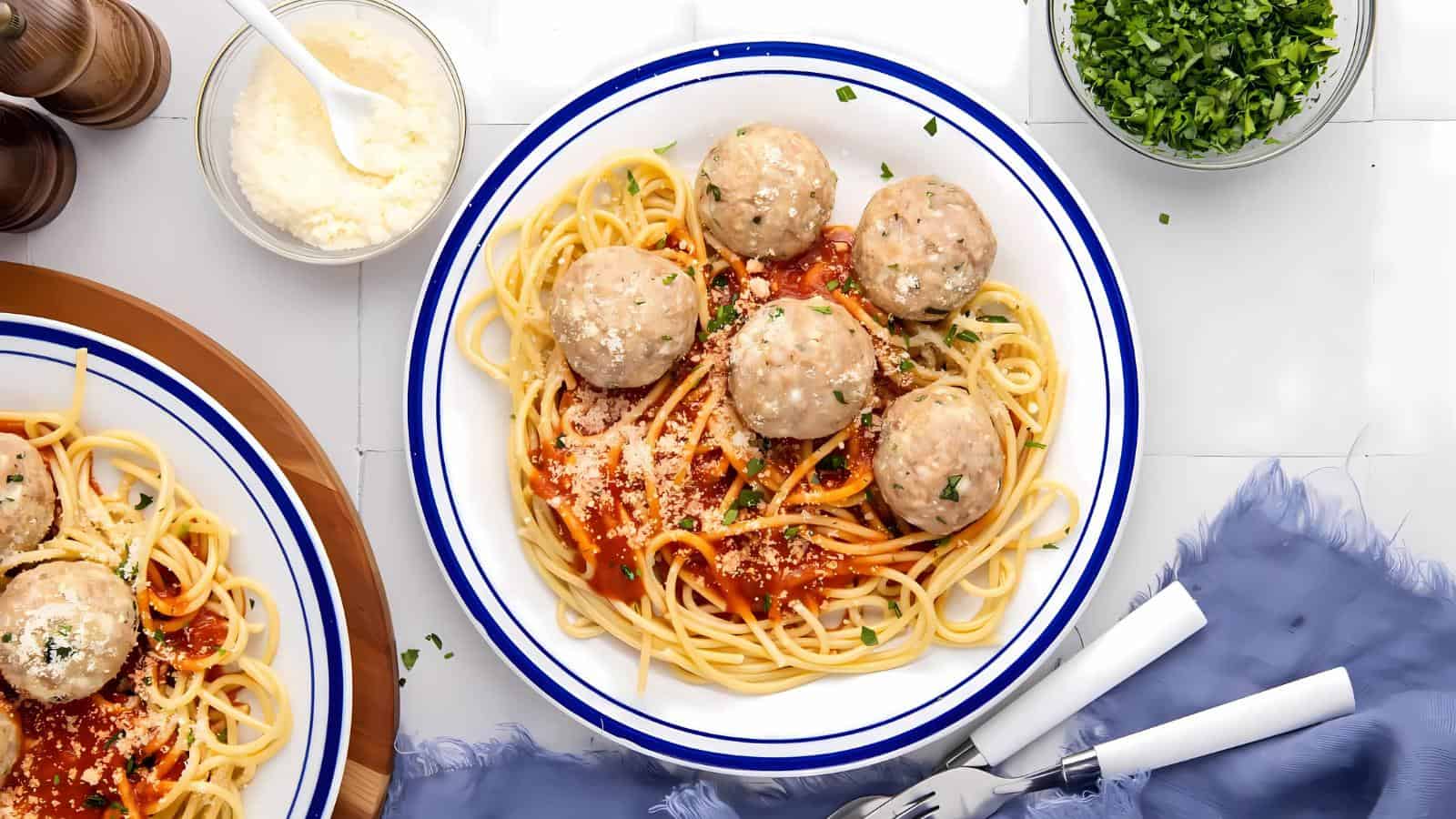 A plate of spaghetti topped with tomato sauce, six meatballs, and grated cheese, garnished with chopped parsley. Bowls of cheese and parsley, utensils, and a napkin are nearby.