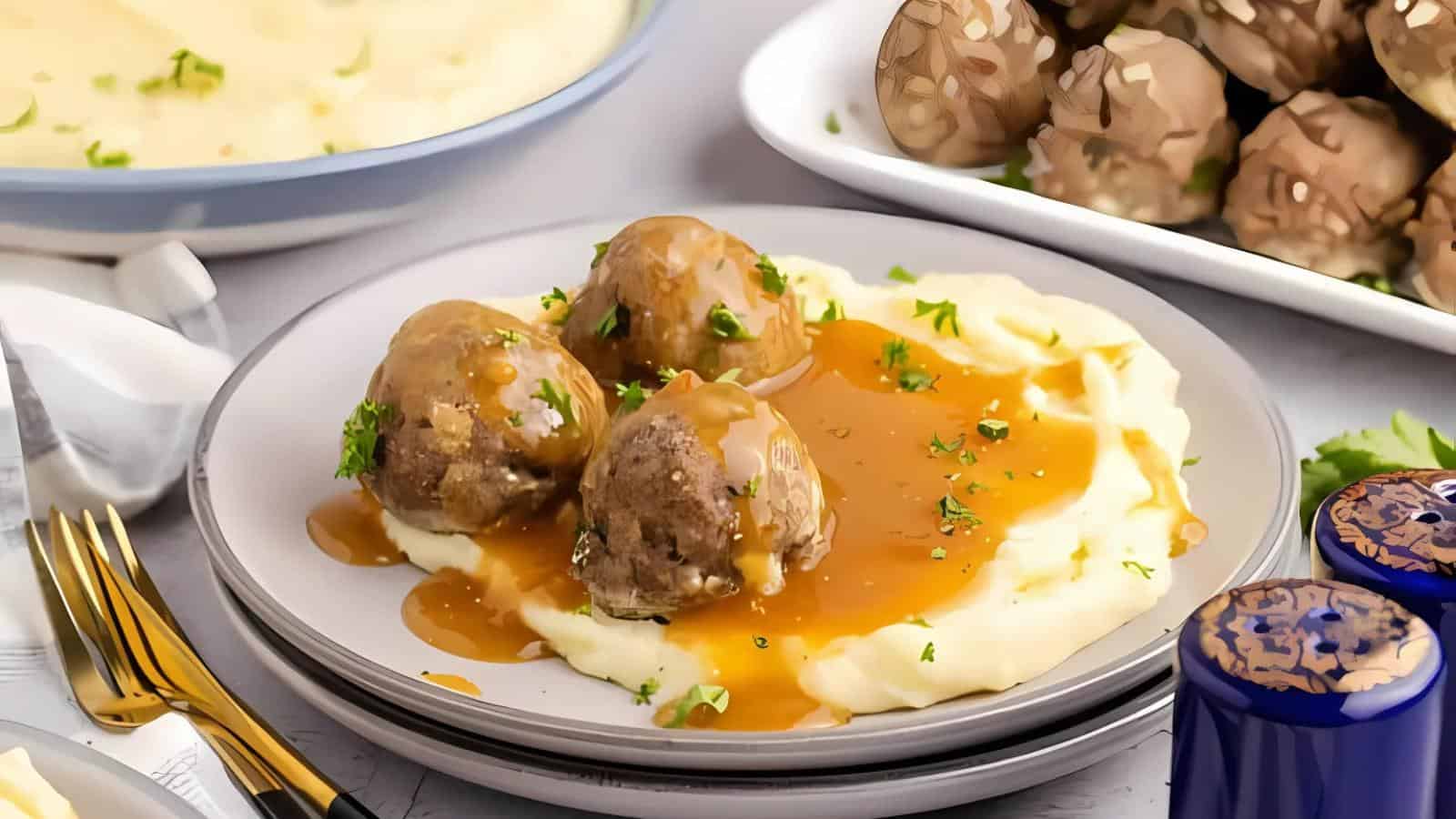 Four meatballs with brown gravy are served on a bed of mashed potatoes, garnished with chopped herbs, with more meatballs and potatoes in the background.