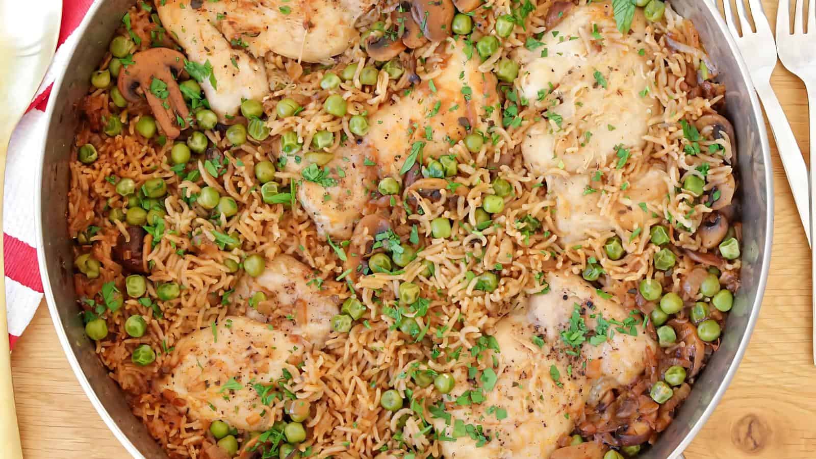A skillet filled with cooked chicken thighs, rice, green peas, mushrooms, and herbs.