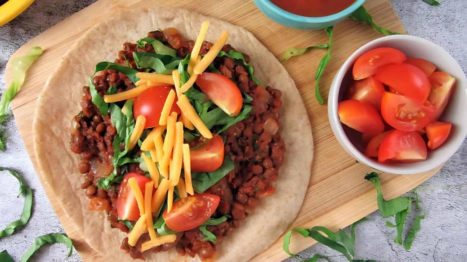 A tortilla topped with cooked lentils, spinach, shredded cheddar cheese, and tomato wedges on a wooden board, with a bowl of tomato wedges nearby.