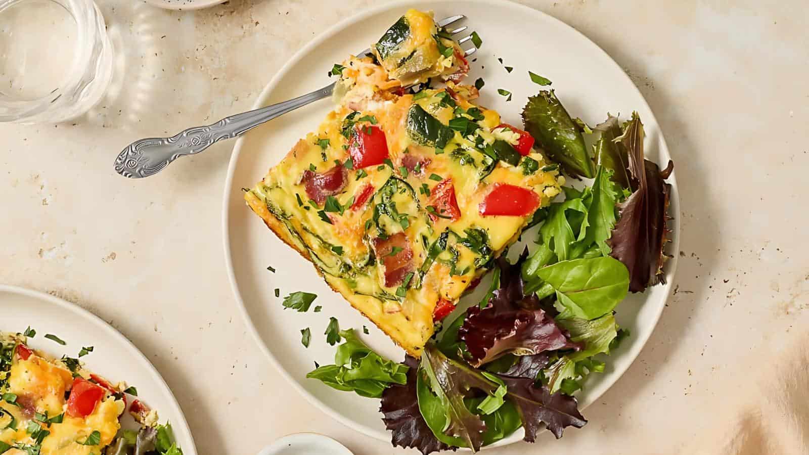 A plate with a serving of vegetable and herb quiche next to a mixed green salad. A fork rests on the plate.