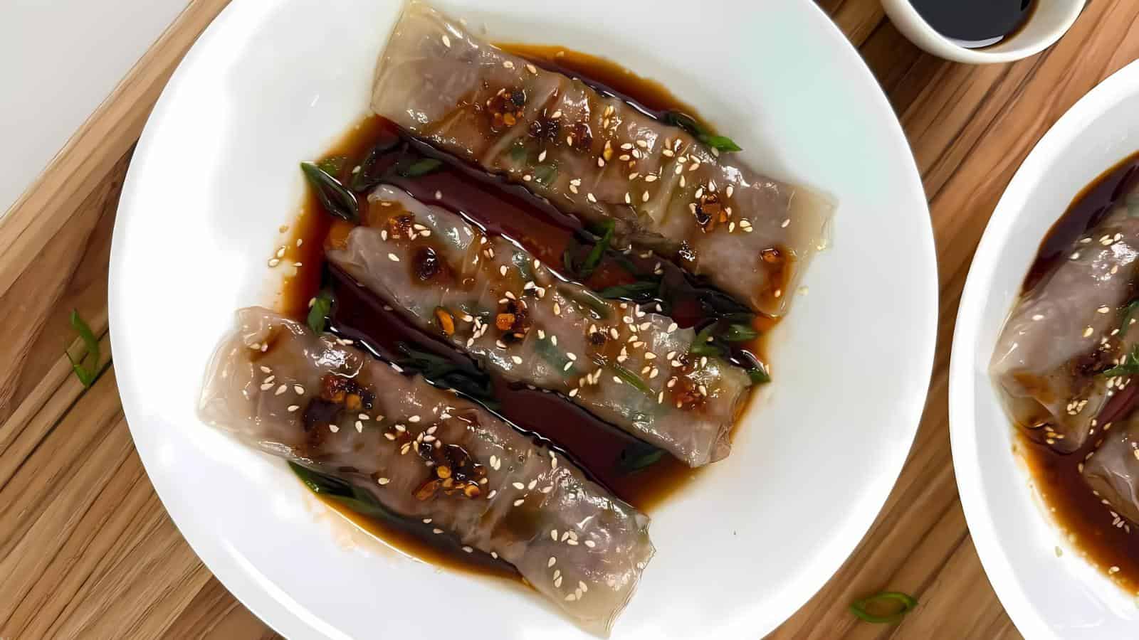 Three translucent rice noodle rolls topped with sauce, sesame seeds, and chopped green onions on a white plate, with soy sauce on the side.