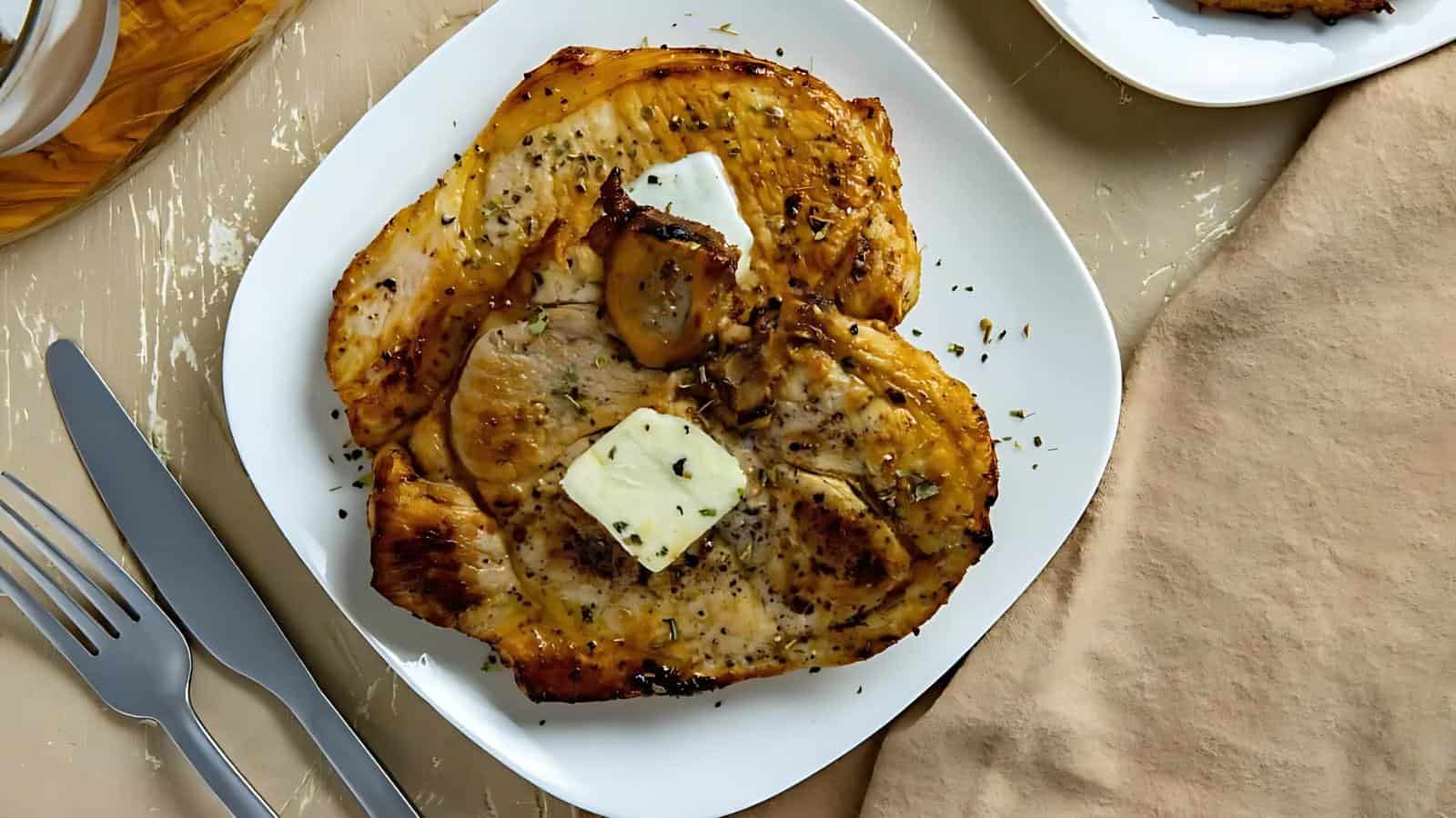 Grilled seasoned chicken breast with two pats of butter on a white square plate, accompanied by a fork and knife on a beige surface.