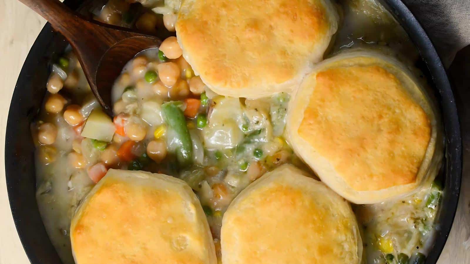 A skillet filled with vegetable pot pie topped with four golden-brown biscuits and a wooden spoon in the mixture.