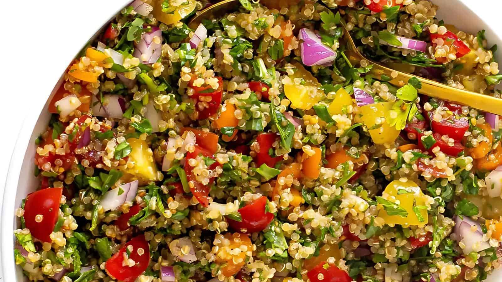 A close-up of a quinoa salad with chopped tomatoes, yellow bell peppers, red onions, parsley, and a gold spoon in a white bowl.