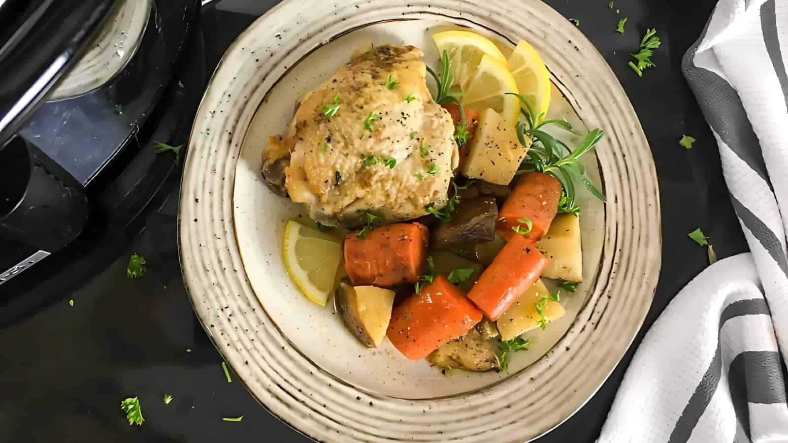 A plate with a cooked chicken thigh, carrots, potatoes, and herbs, garnished with lemon wedges and chopped parsley, sits on a table next to a striped napkin.