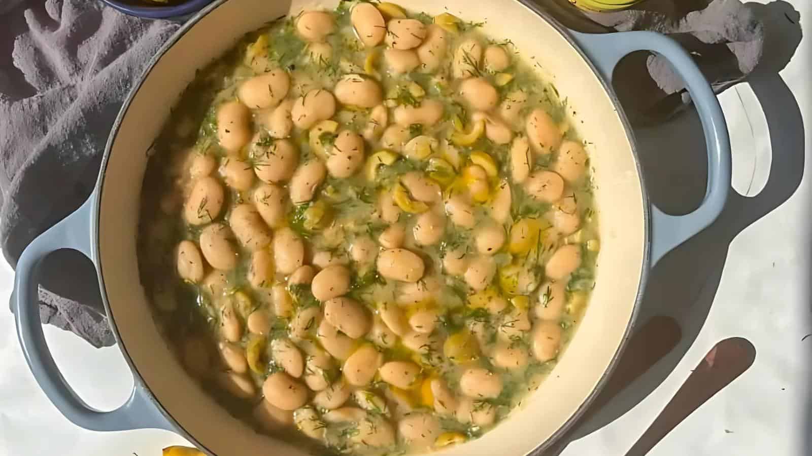 A round blue Dutch oven filled with white beans in a broth with herbs and yellow vegetables, placed on a white surface with a spoon and gray cloth nearby.